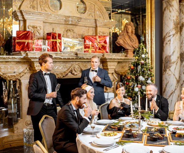 Elegantly dressed group of people having a festive dinner at a well-served table near the fireplace and christmas tree, celebrating New Year holiday at the luxury restaurant